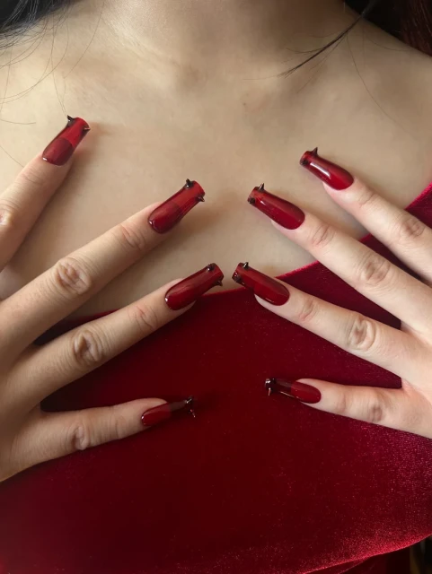 Close-up of a woman's hands with long, red nails featuring small, black spikes, set against a soft, blurred red fabric background.
