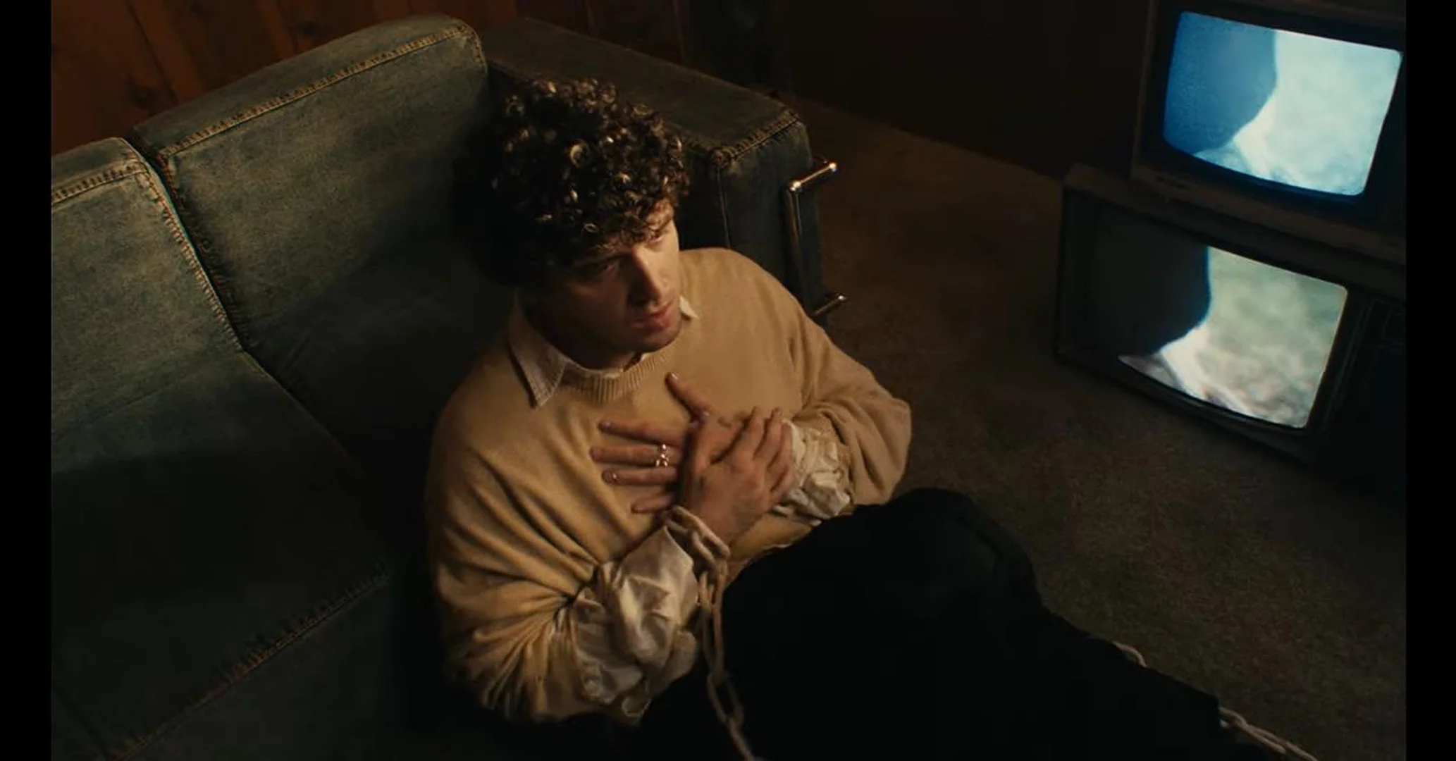 Young man rests on a denim couch in a dimly lit room with two vintage televisions, wearing a beige sweater and black pants.