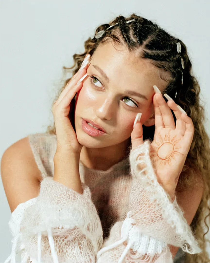 Close-up portrait of a young woman with braided hair and a sheer top, softly lit against a plain background, showcasing beauty and fashion.