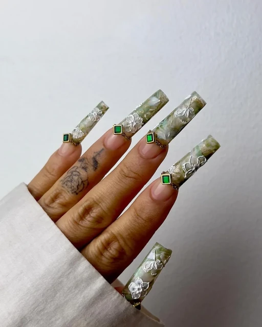 Hand with extra-long nails featuring floral patterns, green gem inlays, and green, gold, and white color palette, against a plain background.