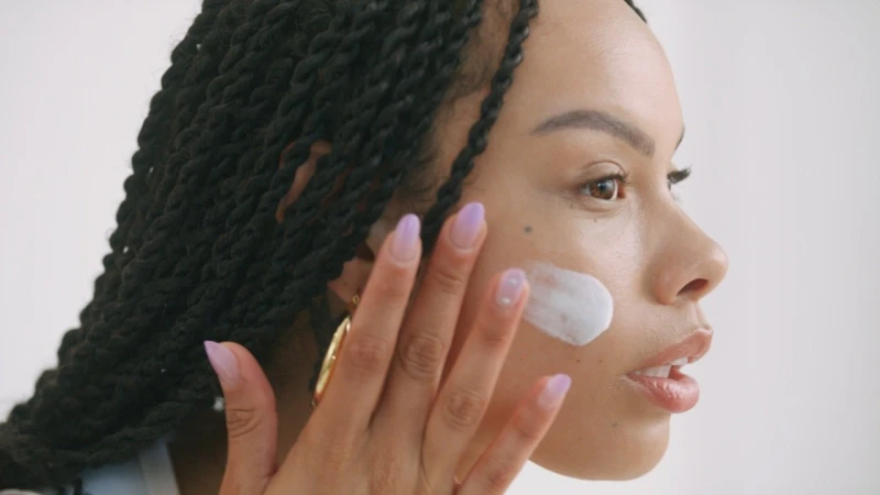 Black woman with braided hair applying white cream to her cheek against a neutral backdrop