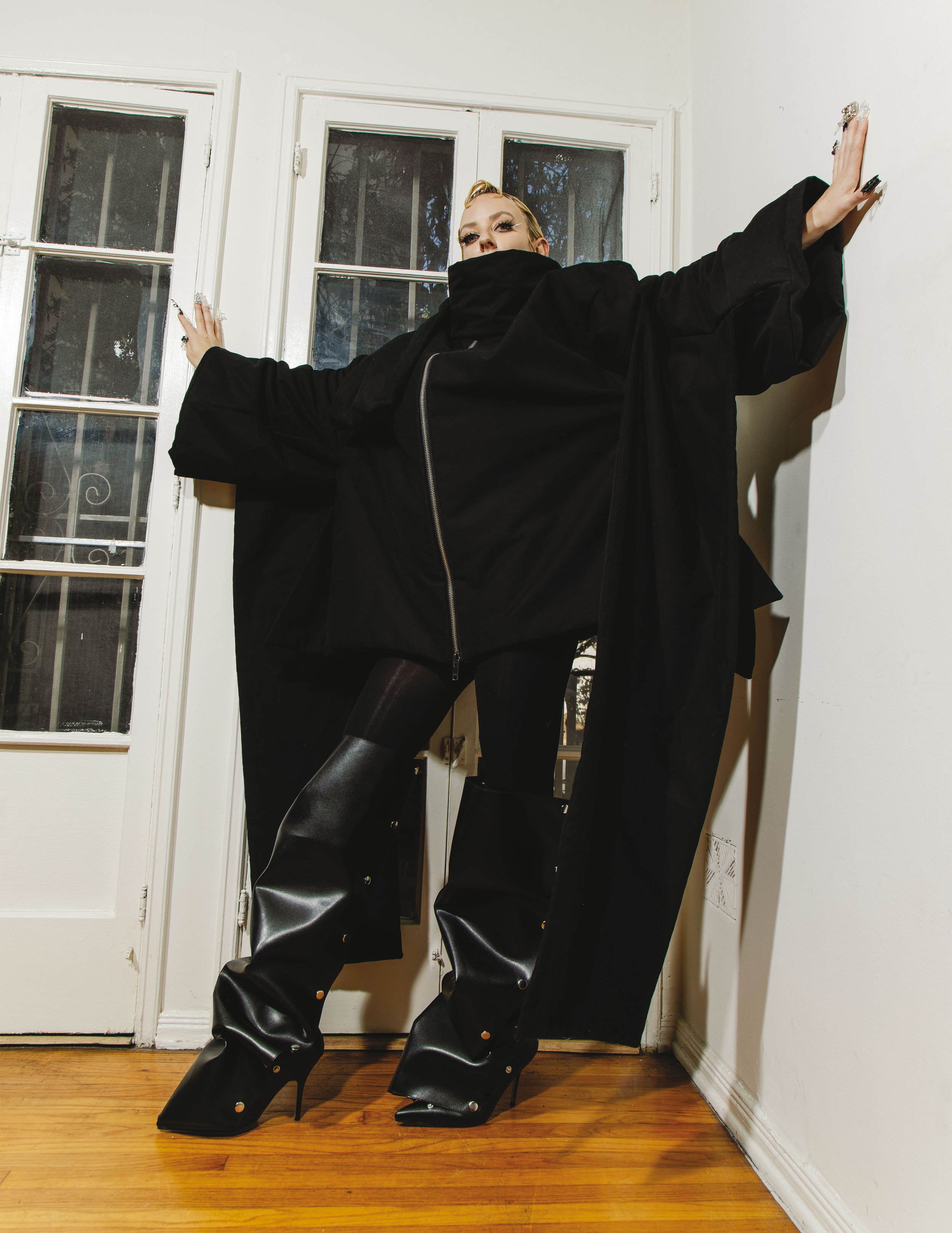 Woman poses in oversized black coat and high heeled boots indoors, with white doors visible in the background.