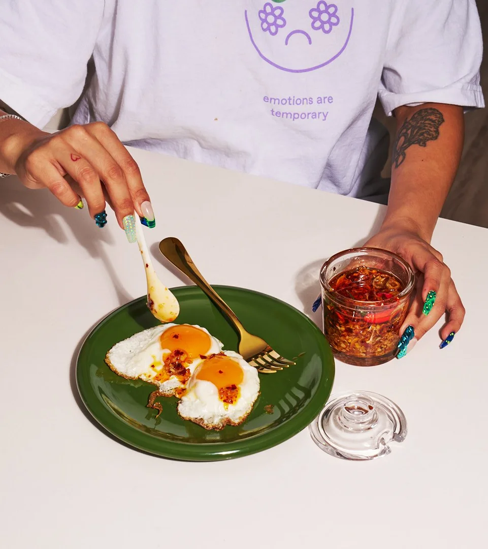 An adult pours chili oil onto fried eggs on a green plate; a jar of chili oil and a white t-shirt with text are also present.