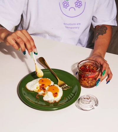 An adult pours chili oil onto fried eggs on a green plate; a jar of chili oil and a white t-shirt with text are also present.