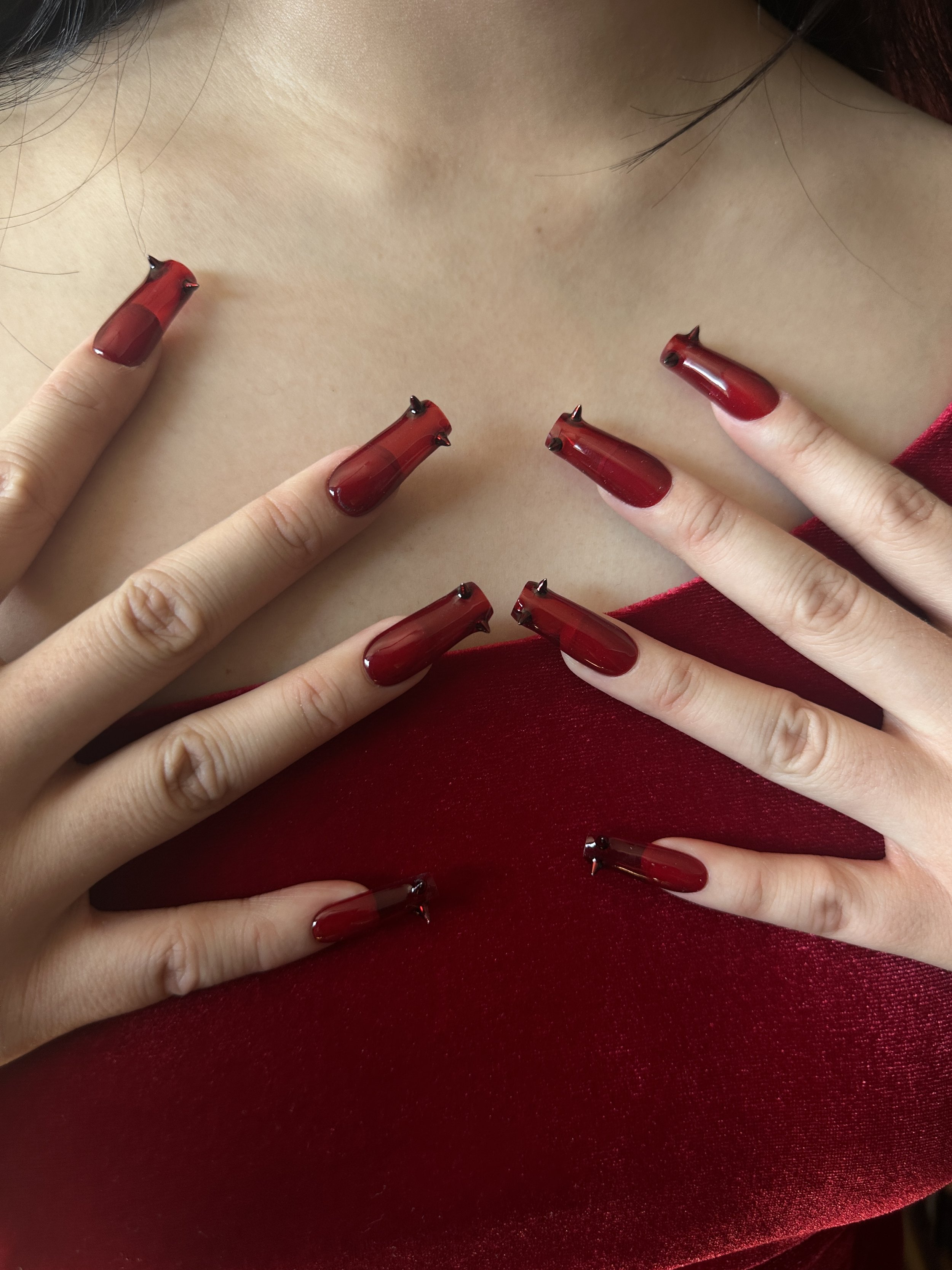 Close-up of hands with red, long, coffin-shaped nails and black nail art details against a matching red velvet top and fair skin.