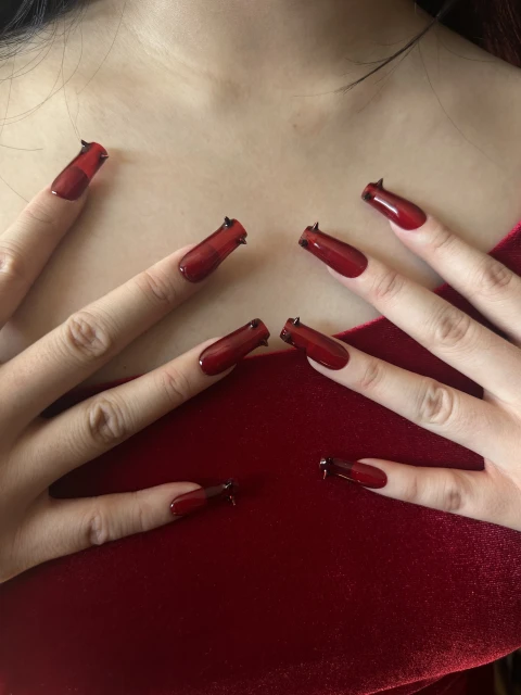 Close-up of hands with red, long, coffin-shaped nails and black nail art details against a matching red velvet top and fair skin.