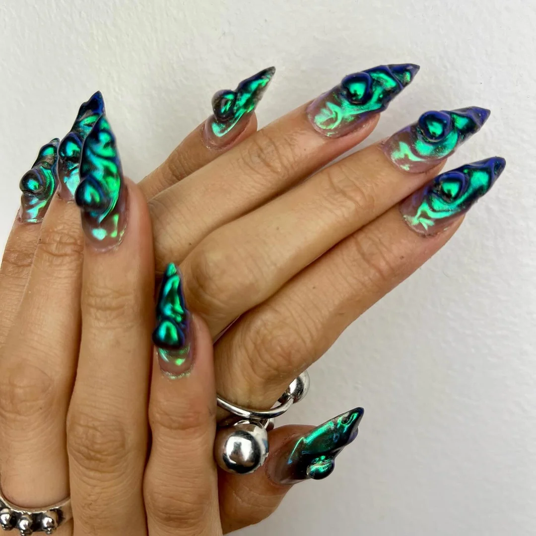 Close-up of hands showcasing long, sculpted nails with a shimmering green and blue design, accented by silver ring details.
