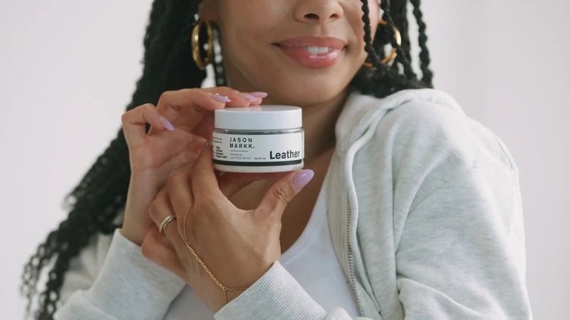Close-up of a smiling woman holding Jason Markk Leather Balm. Indoor shot with a white backdrop, showcasing the product and model.