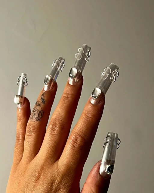 Close-up of a fair-skinned hand with tattoo and long, translucent sculptural acrylic nails adorned with bubble and loop designs.