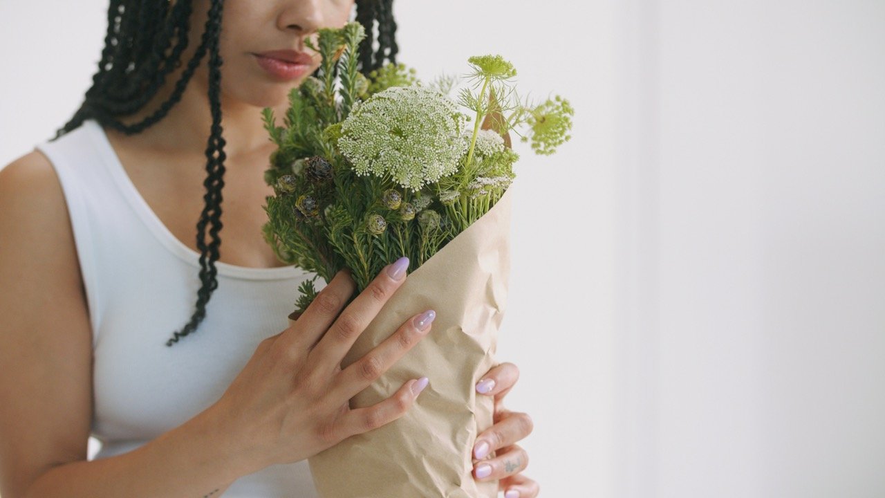 A woman with braids holds a bouquet of wildflowers wrapped in brown paper against a minimalist white background.