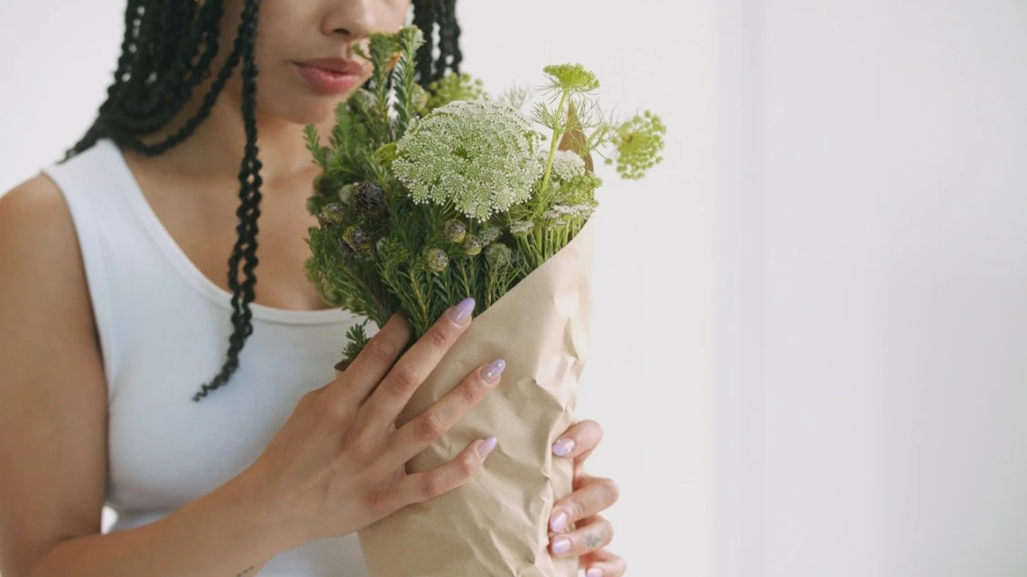 A woman with braids holds a bouquet of wildflowers wrapped in brown paper against a minimalist white background.