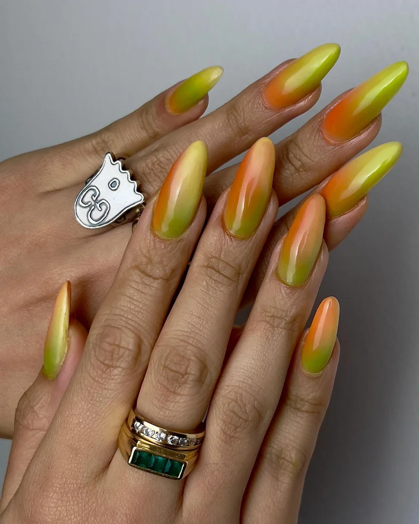 Close-up of hands with long, almond-shaped nails featuring an orange, yellow, and green ombré design, plus silver and gold rings.