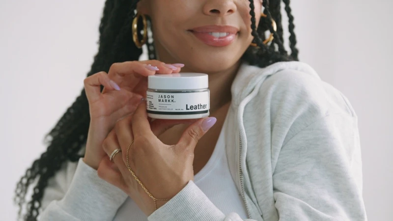 Woman holding Jason Markk leather balm product, wearing gold hoop earrings and a light grey hooded sweatshirt indoors against a white background.