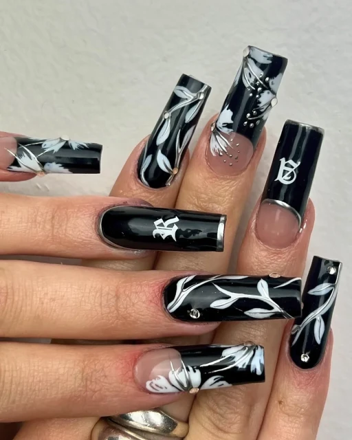 Close-up on hands featuring long square nails adorned with black and white floral designs, accented with silver embellishments, fashion-forward nail art.