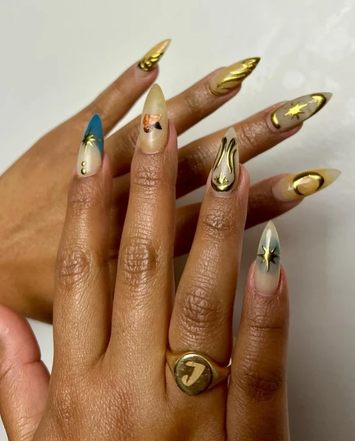 Woman's hands with gold and multi-colored nail art designs, including gold moons, stars, and butterfly, plus a gold initial ring.