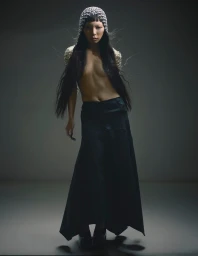 Model with long dark hair wearing a beaded headpiece and dark long skirt against a muted background.