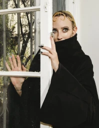 Woman wearing a high-collared black garment looking through a white window frame with decorative nail art.