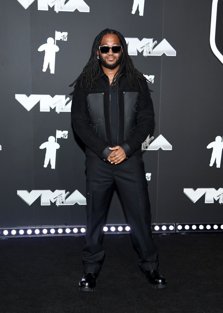 An adult male with dreadlocks and sunglasses stands in front of a VMA-themed backdrop, wearing a black jacket and pants.