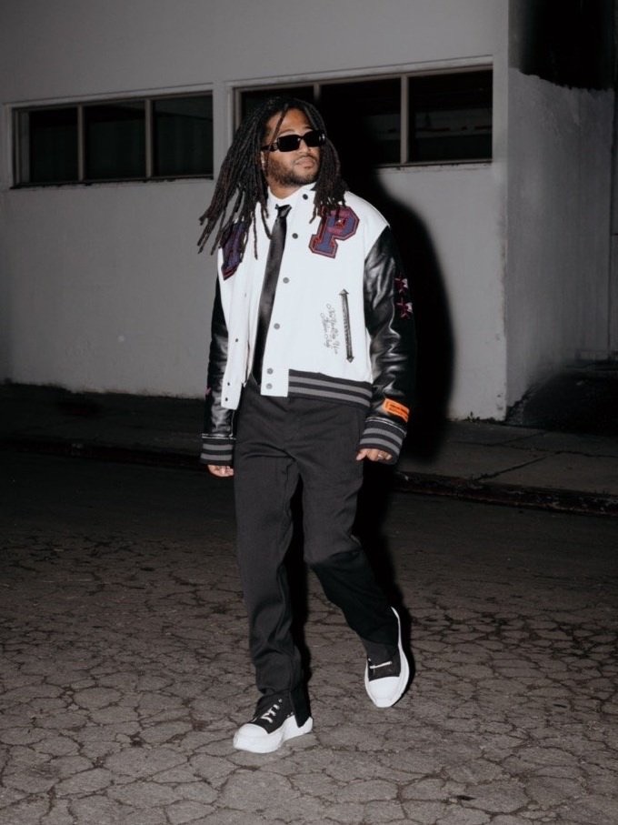 Stylish man in a varsity jacket and sunglasses confidently poses on a city street at night, showcasing urban fashion and modern style.