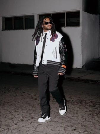 Stylish man in a varsity jacket and sunglasses confidently poses on a city street at night, showcasing urban fashion and modern style.