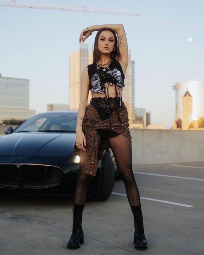 Young woman poses in edgy fashion with a black Maserati on a rooftop against an urban skyline at dusk, showcasing street style.
