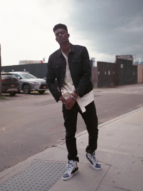 A young African-American man in a denim jacket and sneakers stands confidently on a city sidewalk with parked cars in the background.