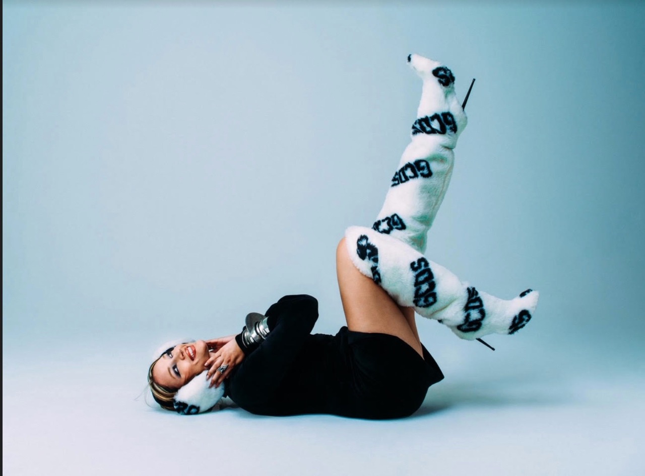 Woman lying on a light blue floor wearing a black dress and furry white boots with black logos poses in a studio.