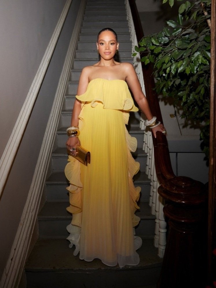A woman stands on stairs wearing a long yellow gown and gold clutch. She is also wearing bracelets, posing and smiling.