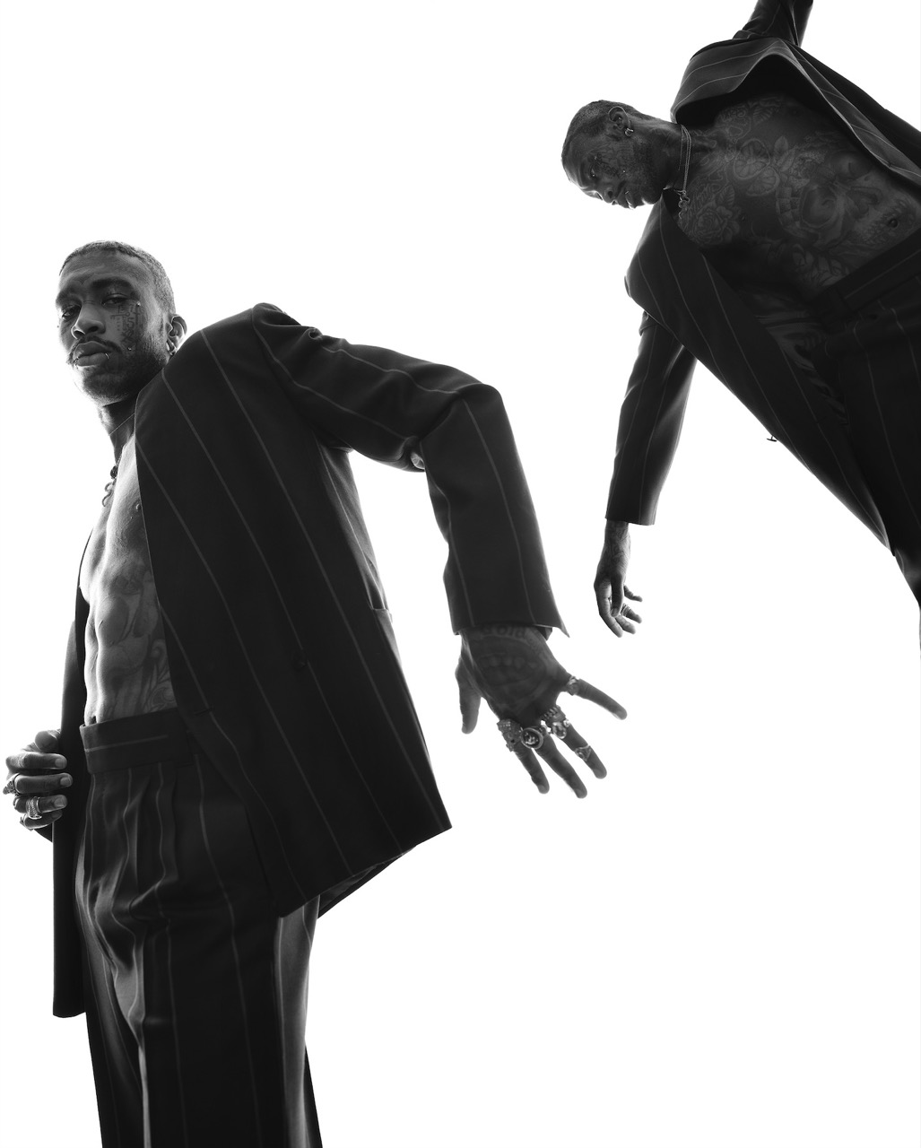 Two African American men strike poses in a studio shot, wearing pinstripe suits, captured from a low angle against a white backdrop.