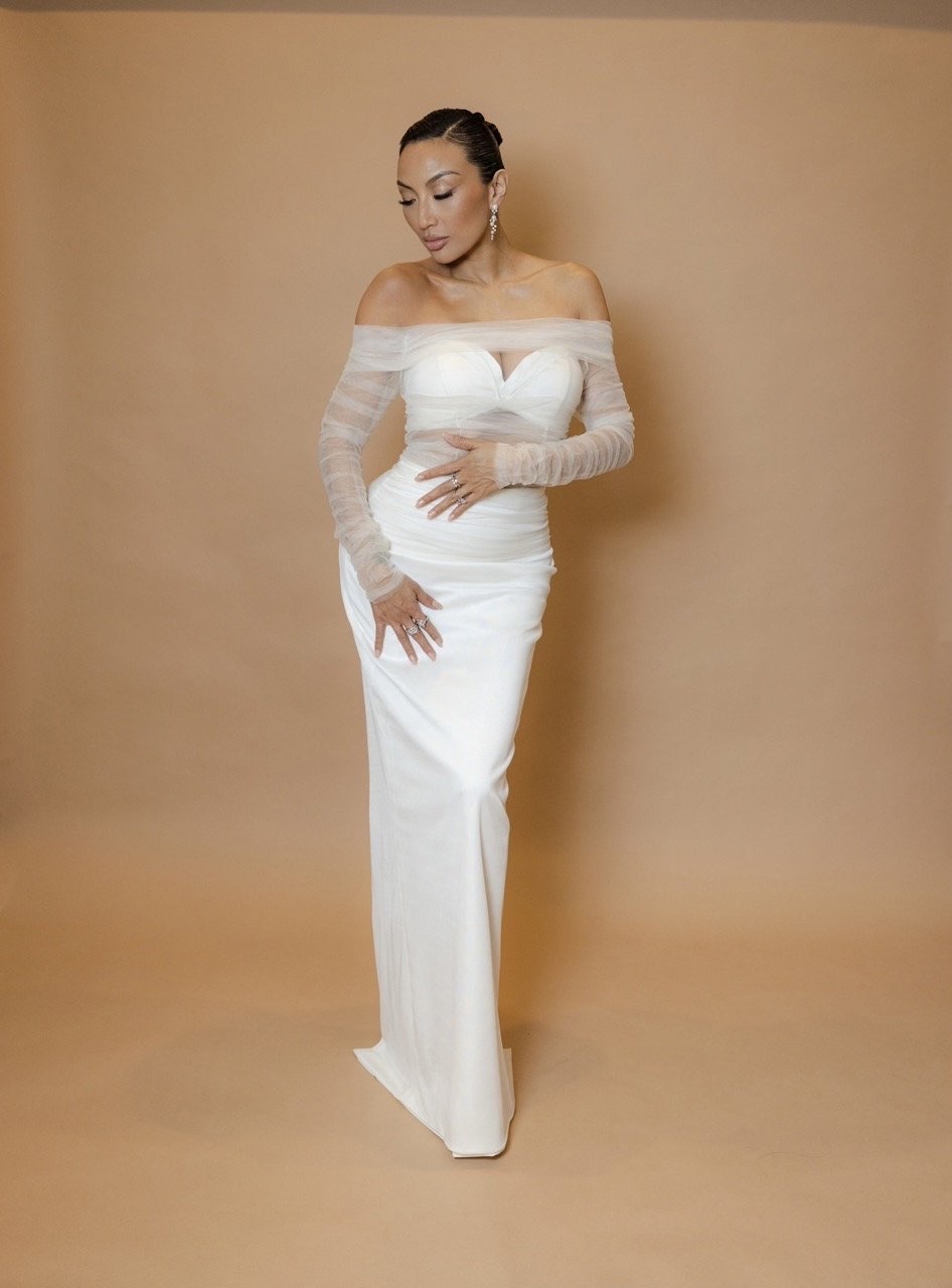Elegant woman in a white off-the-shoulder dress poses against a neutral backdrop, showcasing a refined and classic bridal style.