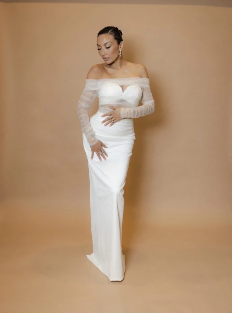 Elegant woman in a white off-the-shoulder dress poses against a neutral backdrop, showcasing a refined and classic bridal style.