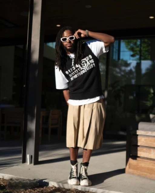 An adult man in a black graphic top, khaki skirt, and high-top sneakers poses outside a modern home with natural lighting.