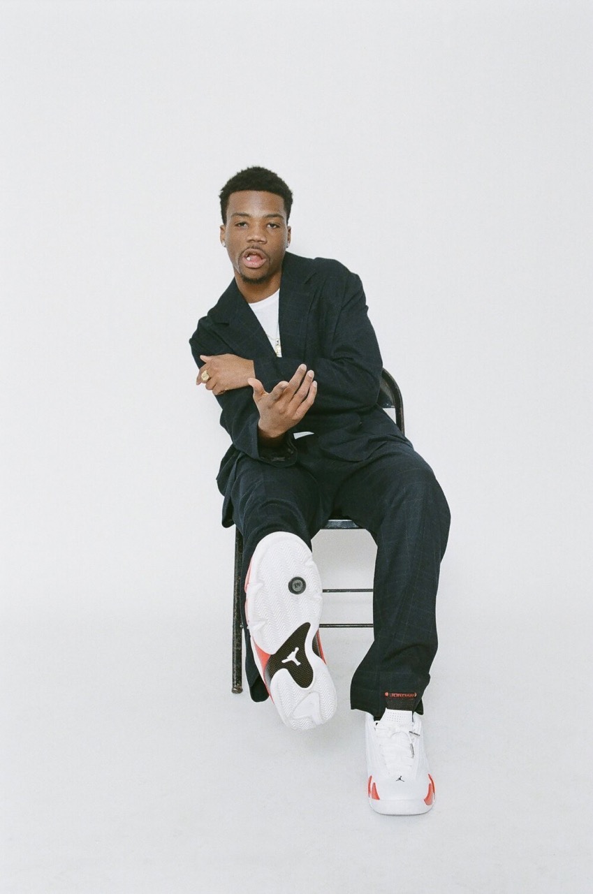 Black man seated on a chair in a dark suit holding one leg forward showing sneakers against a plain white background