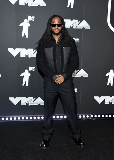 Man in sunglasses, wearing a black jacket and pants, poses against a backdrop featuring the MTV logo and decorative lights.