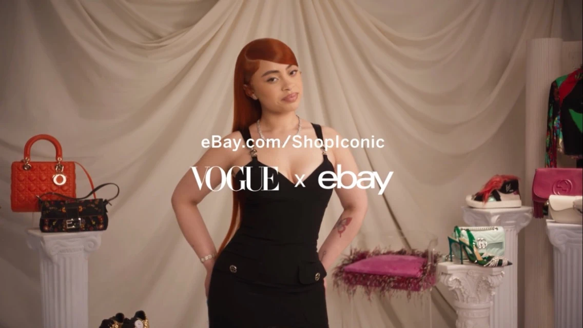 Ice Spice poses in a black dress in front of a cream drape. Fashion bags and shoes are displayed on pillar stands.