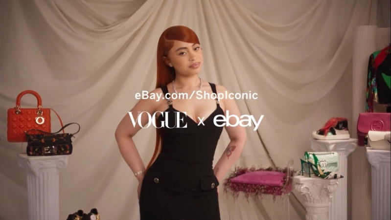 Ice Spice poses in a black dress in front of a cream drape. Fashion bags and shoes are displayed on pillar stands.