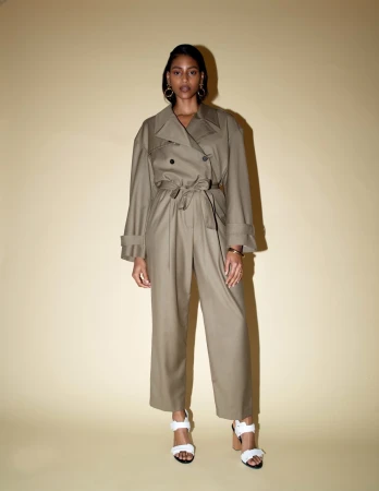 Model in olive green belted trench jumpsuit and white heeled sandals posing against a warm neutral studio backdrop.