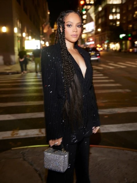 Adult female in a black sequined suit stands on a city street at night with dark hair braided, holding a sparkling box bag.