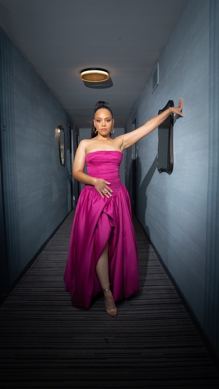 Elegant woman in a pink strapless gown stands in an indoor hallway with gray walls, creating a striking contrast and fashion portrait.