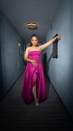 Elegant woman in a pink strapless gown stands in an indoor hallway with gray walls, creating a striking contrast and fashion portrait.