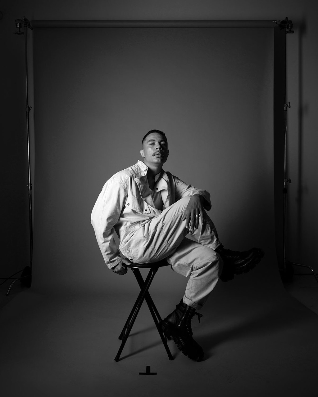 Black and white studio portrait of a stylish adult male in a jumpsuit, sitting on a stool, with backdrop and lighting stands.
