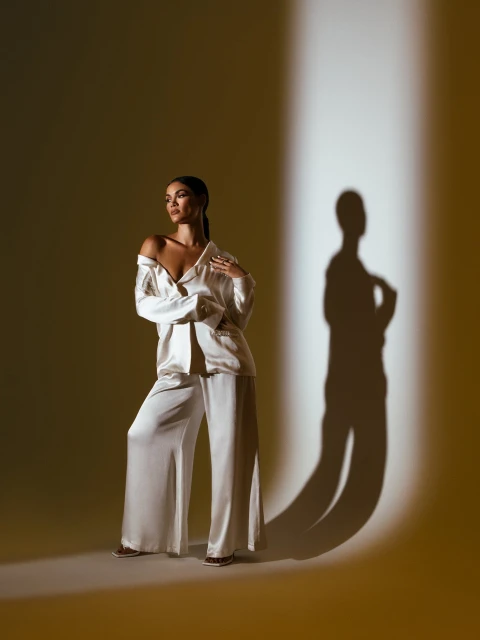Full-length shot of a woman in a white satin pajama set in a studio, with a shadow cast on the wall and warm lighting.