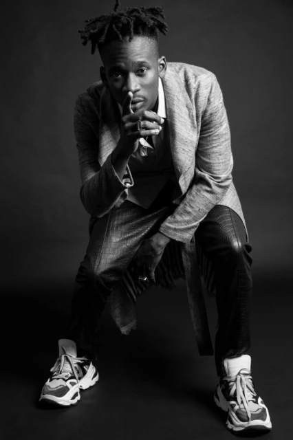 Black and white portrait of a Black man with dreadlocks wearing a grey suit and sneakers, posing with finger to lips in a photo studio.