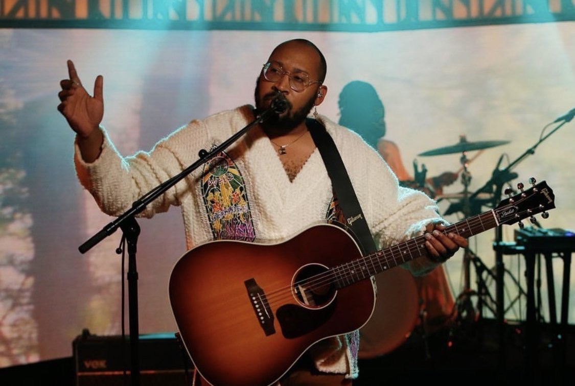 Singer-songwriter performing live with an acoustic guitar, wearing a patterned sweater and glasses, on a concert stage with warm lighting.