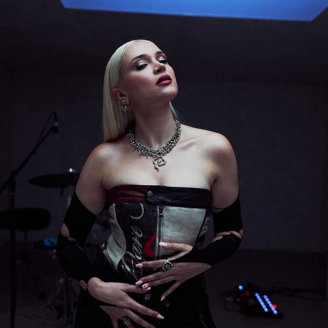 Portrait of an adult female with blonde hair wearing a strapless top and silver jewelry in a dimly lit indoor environment.