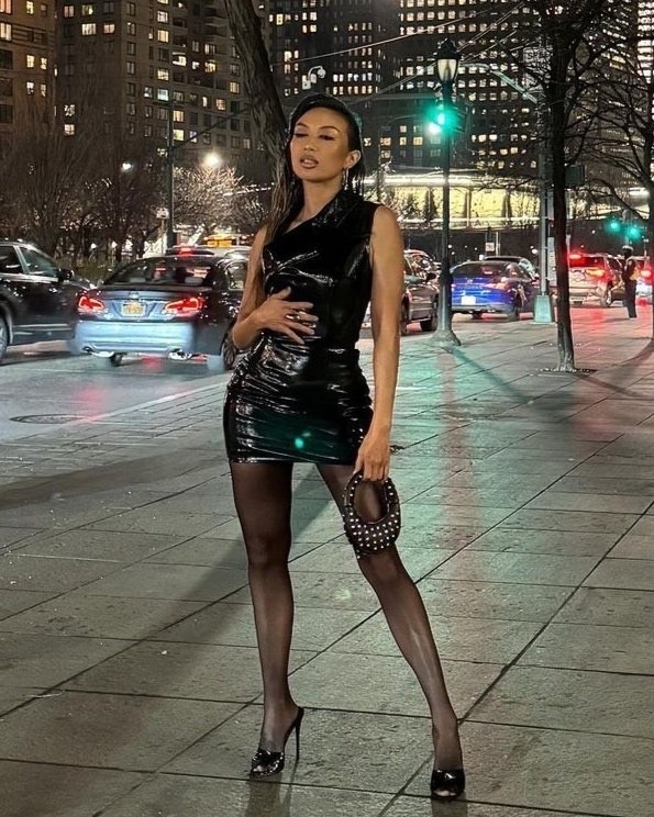 Adult female in a black latex dress stands on a sidewalk at night in an urban environment. Cars and illuminated buildings in the background.