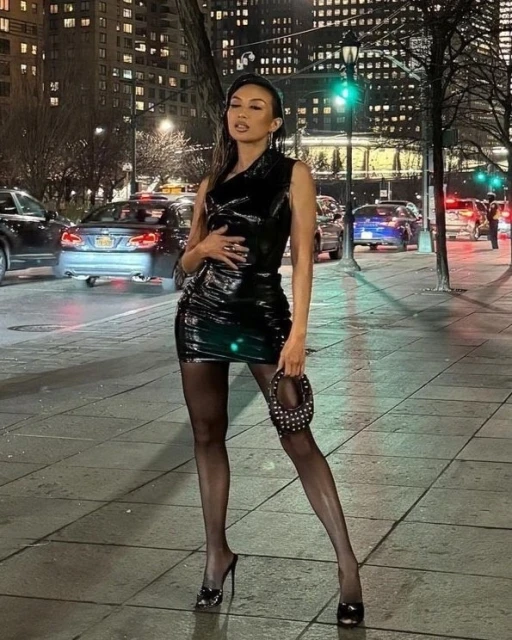 Adult female in a black latex dress stands on a sidewalk at night in an urban environment. Cars and illuminated buildings in the background.