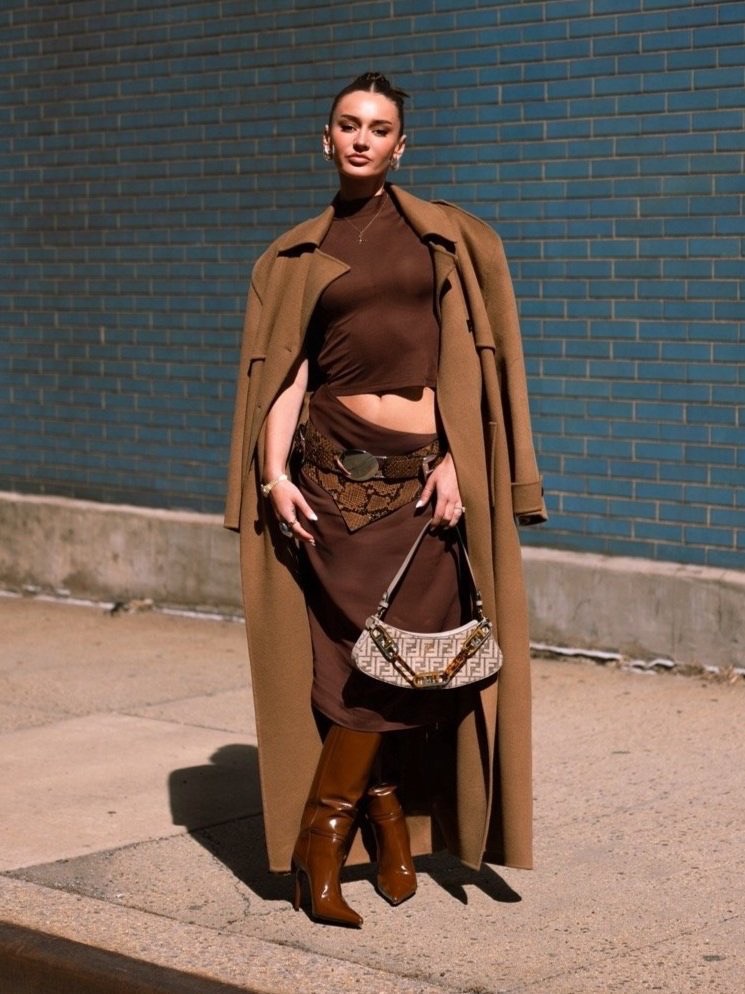 A woman stands outdoors against a blue brick wall, wearing a brown top and skirt, tan overcoat, boots and carrying a small purse.