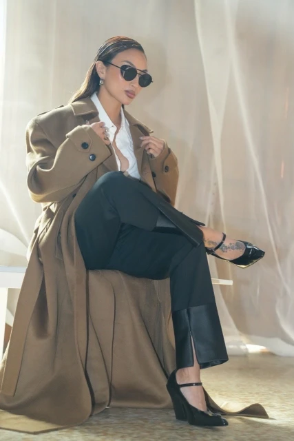 Elegant woman in a tan trench coat, black pants, and sunglasses sitting indoors with sheer curtains in the background; fashion studio shot.