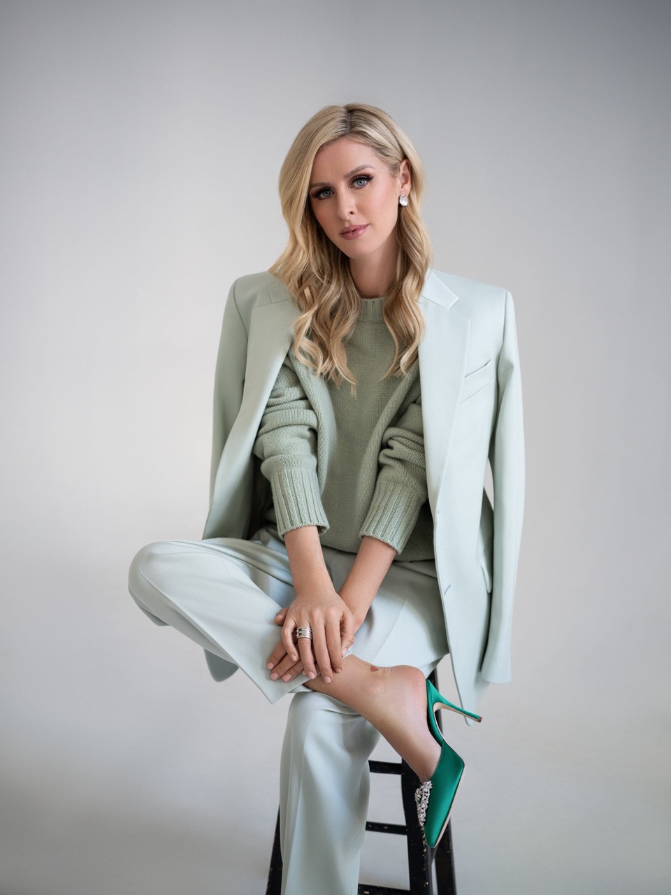 Blonde woman in light green suit poses seated on a stool in a studio setting, showcasing a modern and elegant fashion aesthetic.